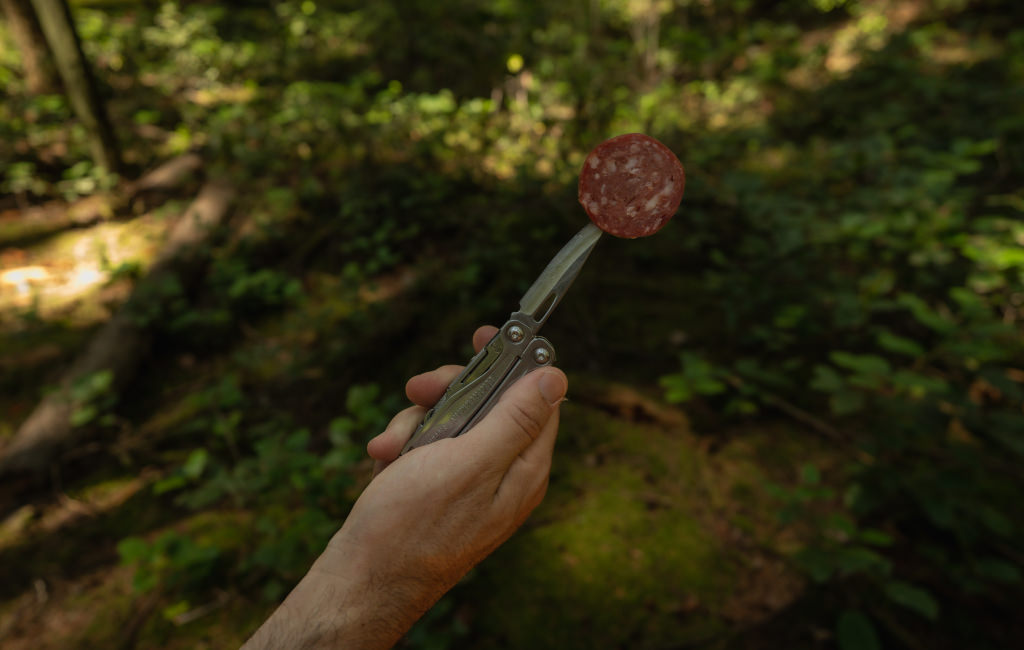 A person stands in the woods, gripping a Leatherman Sidekick with meat on knife blade, surrounded by tall trees and dappled sunlight filtering through the leaves.