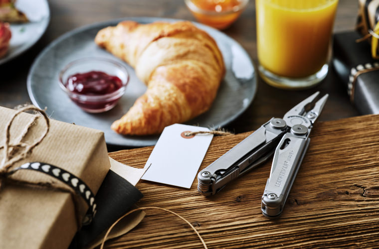 A table set with fresh croissants, a glass of orange juice, Leatherman Wave+ and a decorative gift tag for a delightful breakfast scene.